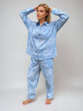 Load image into Gallery viewer, A woman modeling a light blue pajama set with a white floral print, consisting of a long-sleeve button-up shirt and matching pants, against a neutral gray studio background.