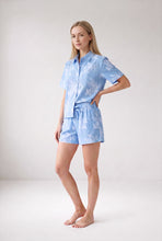 Load image into Gallery viewer, A woman modeling a light blue short-sleeve button-up shirt and matching shorts set with a white floral pattern, standing against a plain light gray studio background.