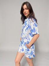 Load image into Gallery viewer, A woman modeling a white short-sleeve pyjama set in organic cotton with a blue floral pattern, standing against a neutral gray studio background.