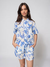 Load image into Gallery viewer, A woman modeling a matching short-sleeve collared shirt and shorts set with a blue floral print on a white background, standing against a light gray studio backdrop.