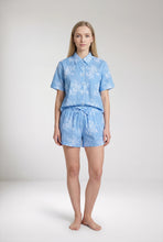 Load image into Gallery viewer, A woman stands barefoot against a light gray studio background wearing a light blue short-sleeve collared shirt and matching shorts with a white floral pattern.

