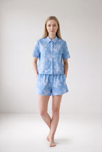 Load image into Gallery viewer, A woman modeling a light blue short-sleeve button-up shirt and matching shorts set with a white floral pattern, standing against a neutral light gray studio background.
